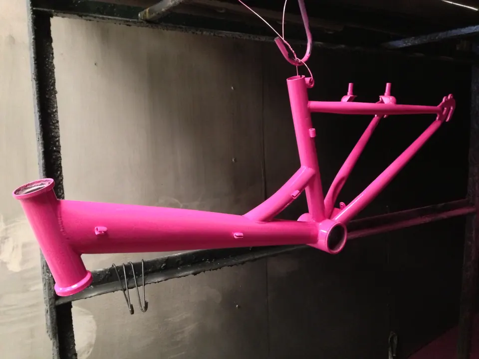 A child's bike frame powder coated bright pink, hanging in the powder coating booth